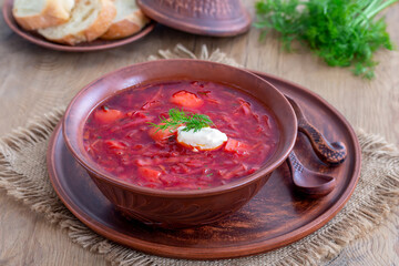 Eastern European Borscht served in earthenware. Red beetroot soup topped with a scoop of sour cream and fresh sprigs of dill on the wooden table. Close-up, selective focus.