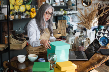 Attractive aged woman in eyeglasses and apron doing inventory at decor store. Competent saleswoman rechecking all available assortment at shop.
