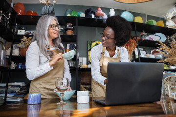 Two saleswomen of different age and race counting goods at decor shop using modern laptop. Concept of people, technology and inventory.