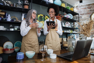 Young african woman and mature caucasian woman standing at decor shop and using digital tablet for receiving goods. Working process at store.