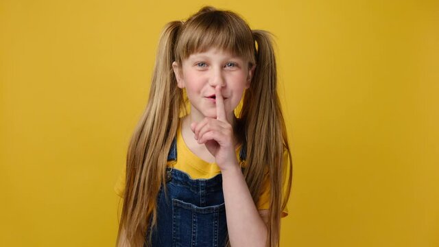 Portrait of secret little schoolgirl kid keeps finger on lips, shows silence sign, asks to be quiet, not to make loud noise. Attractive cute child model poses against yellow studio background wall
