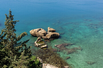 View of pure blue sea water, huge stones and tops of green trees.