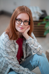 A close-up portrait of a young girl in glasses and casual clothes sitting on the floor of her new apartment