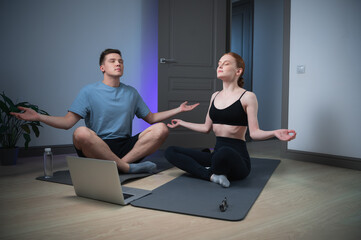 Obraz premium A husband and wife spend a yoga class at home sitting on the floor, they meditate together in front of a laptop