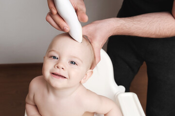 First haircut of smiling toddler baby. Young father cuts first hair of his son indoors. Man's hands shave a little boy's head with electric hair clipper. 