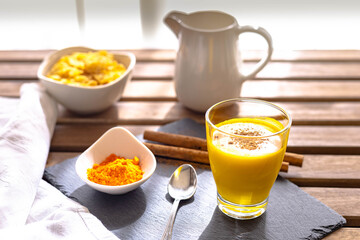 selective focus of a breakfast of golden milk with curcuma and cinnamon in a glass placed on a slate board and cornflakes in the background on a table with the light from the window