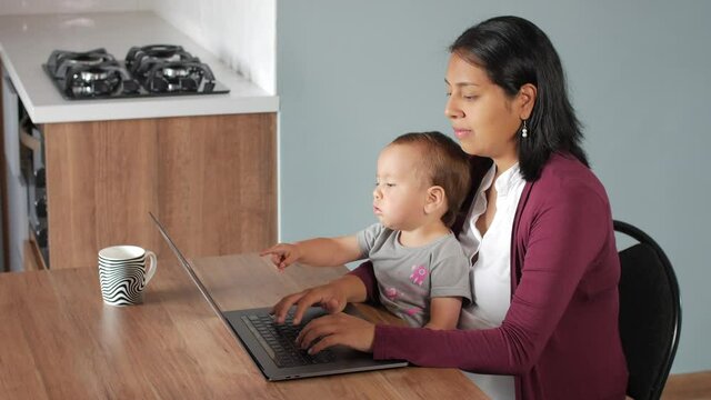 A Hispanic mother is sitting working or studying using a laptop at a table together with baby girl 