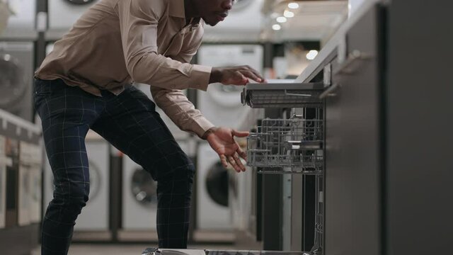 shopping in home appliance store in department with dishwasher, young black guy is viewing modern device for kitchen, automatic dish washer