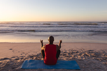 Rear view of senior african american man meditating and practicing yoga while sitting on yoga mat at - Powered by Adobe