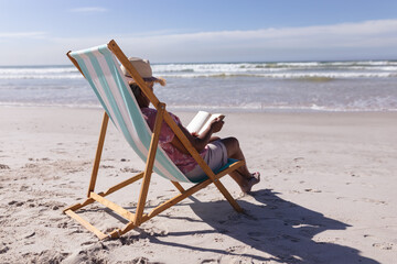 Senior african american woman reading a book while sitting on deck chair at the beach