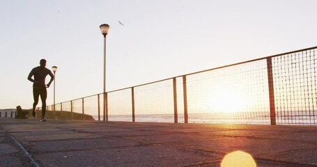 Focused african american man running exercising outdoors by seaside at sunset