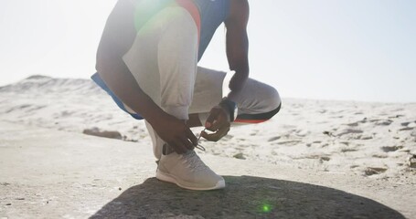 Midsection of african american man tying shoelaces during exercise outdoors on beach - Powered by Adobe