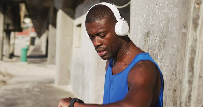 African american man wearing headphones checking his smartphone, exercising outdoors on sunny day