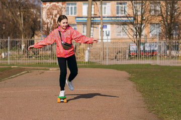 Obraz premium Happy smiling teenage girl in bright clothes ride a skateboard or penny board in the park. Active sports recreation that develops coordination. Free space for text