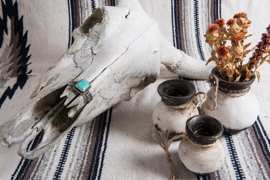 Weathered cow skull, turquoise and silver cuff bracelet, black and white painted Mexican pottery with dried flowers on a Mexican striped blanket.