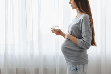 Young beautiful pregnant woman standing at home near the window and drinking water with lemon.