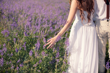 Newlyweds are walking along summer field. Bride touches wildflowers with her hand. Elopement wedding. Selective focus.