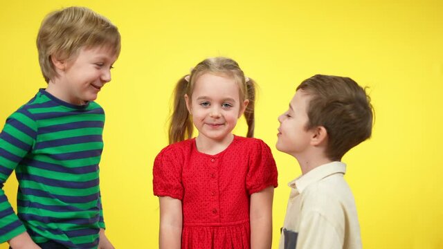 Two boys kissing pretty little girl at yellow background. Portrait of playful smiling children having fun. Brothers and sister or friends together. Childhood and happiness concept