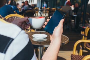 Man holds a smartphone on the background of a cup of coffee in a restaurant.