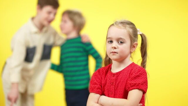 Sad charming little girl with crossed hands looking at camera as blurred boys mocking at yellow background. Portrait of upset child posing with bullied pointing and laughing. Bullying and sadness