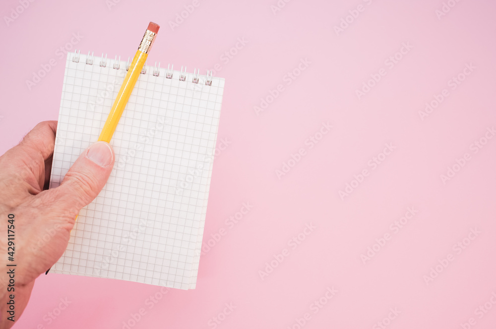 Poster Closeup shot of a hand holding a pencil with a notebook on a pink backgrou