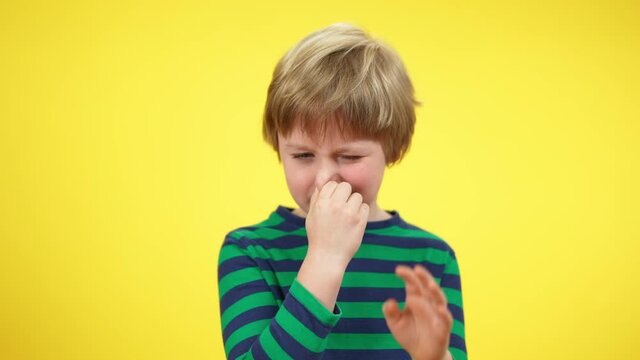 Cute blond little boy closing nose smelling unpleasant smell. Portrait of charming Caucasian child posing at yellow background. Disgust and bad scent