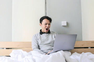 Surprised and shocked Asian man is working with his laptop on his cozy bed. Concept of successful freelancer lifestyle.