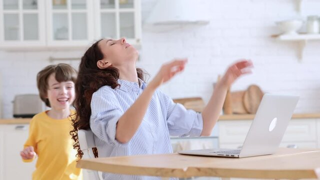 Exhausted mom sits by table at home office during lockdown working on laptop trying to concentrate. Noisy child distracts from work pulls the sleeve and hair asking attention from annoyed busy mother 