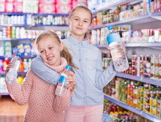 Portrait of cheerful little girls standing among shelves in a supermarket, holding a bottled water