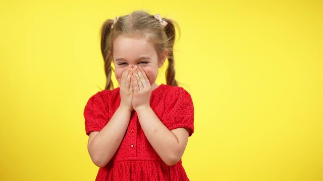 Funny cute little shy girl laughing closing mouth with hands. Portrait of beautiful blond Caucasian child with grey eyes posing at yellow background having fun