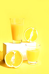 Fototapeta premium A glass of freshly squeezed refreshing orange juice on a white pedestal or podium with orange halves. Yellow background. The concept of a healthy lifestyle, raw food diet, vegetarianism. Copy space