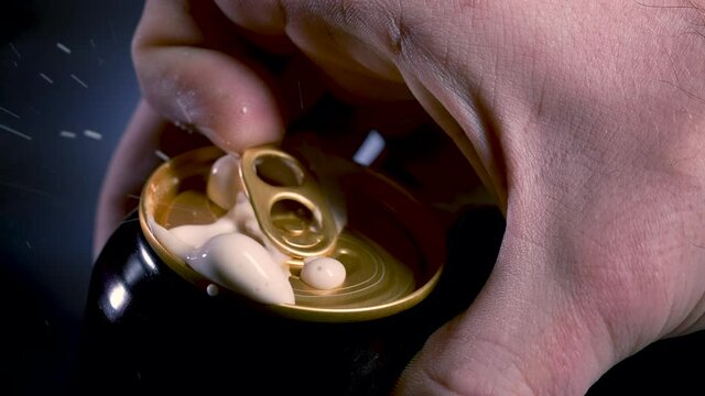 Opening a shaken can of beer, from which the foam flows. A detailed shot of a man's hand opening a stirred beer can against a black gradient background. Slow motion. Close up.