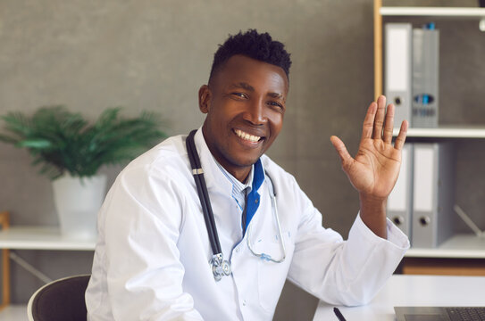 Headshot of happy young African-American doctor saying hello to patients. Portrait of handsome black man in white lab coat looking at camera, smiling and waving hand