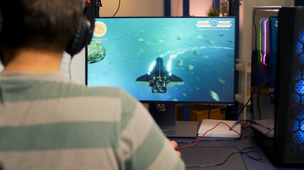 Over shoulder footage of professional streamer playing digital space shooter video games on computer using headphones, microphone and mouse. Videogamer man streaming online video games in room with