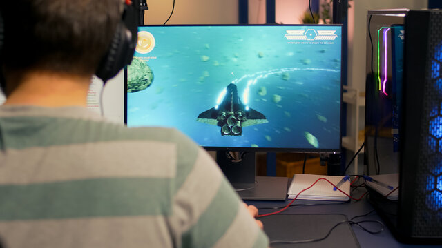 Over shoulder footage of professional streamer playing digital space shooter video games on computer using headphones, microphone and mouse. Videogamer man streaming online video games in room with - Powered by Adobe