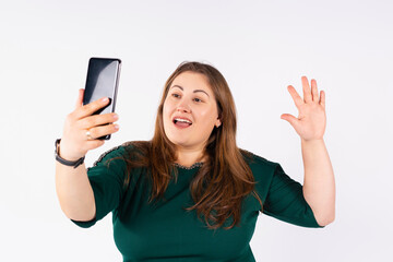 Fototapeta premium Portrait of a happy caucasian plump american woman in retro style clothes, green blouse waving standing and taking a selfie on modern smartphone isolated over grey background. Overweight happy people