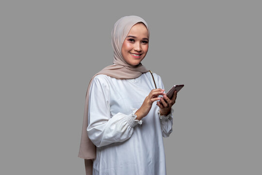 Muslim woman in hijab is holding a cellphone smiling and looking at the camera isolated on plain background