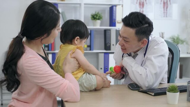 friendly male physician is playing with the child patient by not letting her touch the handheld rattle on purpose during a routine visit in his office