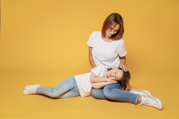 Fototapeta na wymiar Full body length happy woman in basic white t-shirt sit on floor child baby girl 5-6 years old sleep. Mom mum little kid daughter isolated on yellow color background studio. Mother's Day love family.