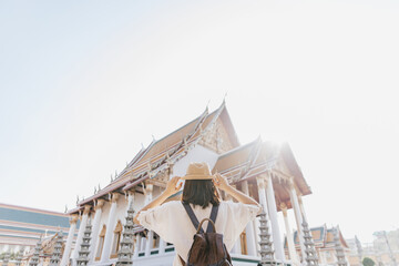 Behind of tourist woman in white shirt and straw hat with brown backpack looking at Wat Suthat Thepwararam Ratchaworamahawihan Temple, Bangkok, Thailand