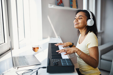 Joyful young woman playing synthesizer and using notebook at home