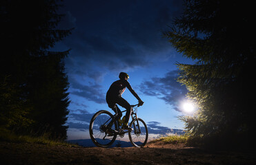 Back view of man cyclist riding bike under blue evening sky with clouds. Silhouette of bicyclist riding bicycle on the trail in night mountain forest. Concept of sport, biking and active leisure.