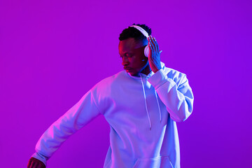 Young handsome African man wearing headphones listening to music and dancing in futuristic purple...