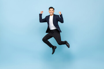 Fototapeta na wymiar Full lenght portrait of smiling handsome Asian man jumping and raising his fists on isolated light blue studio background