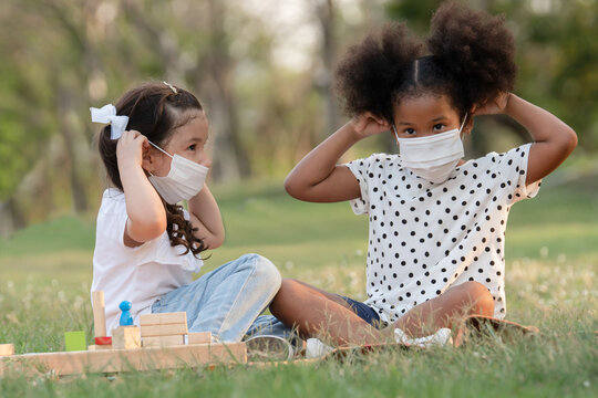 Couple little girls African and Caucasian kids wearing face mask while sitting and playing wooden blocks toy in green park together. Ethnic diversity of friendship in new normal lifestyle concept