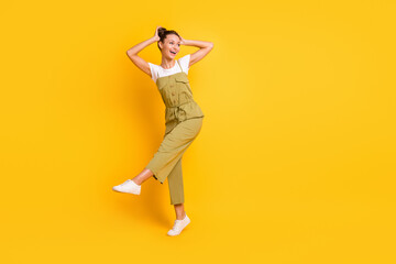 Full size photo of optimistic brunette lady dance look empty space wear bright t-shirt overall sneakers isolated on yellow color background