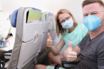 Fototapeta na wymiar Man and woman in protective medical masks flying in airplane and showing thumbs up