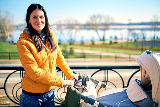 A happy young mother keeps a stroller with a child on the street to the park and looks into the camera. outdoor leisure. Sunny weather and beautiful scenery. Healthy lifestyle