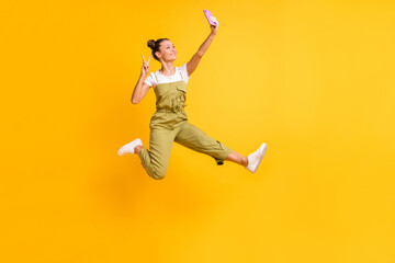 Fototapeta na wymiar Photo of funky lady jump hold phone make selfie show v-sign wear green overall sneakers isolated yellow background