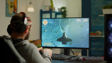 Pro man gamer playing video game at powerful personal computer late night wearing professional headphones. Excited player using wireless controller for virtual tournament gaming space shooter at home © DC Studio